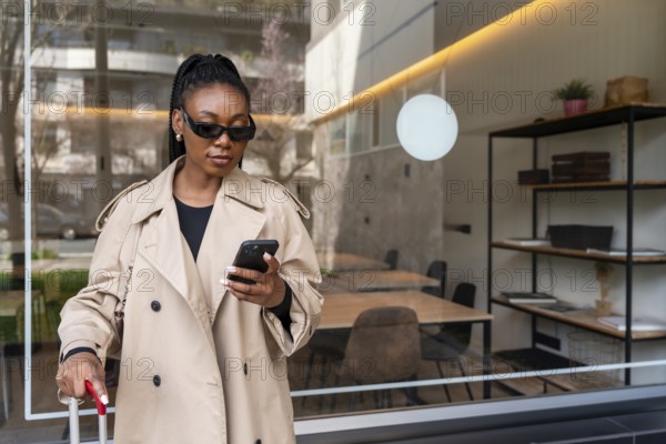 Stylish businesswoman using smartphone while holding luggage outside hotel during business trip, epitomizing modern work travel
