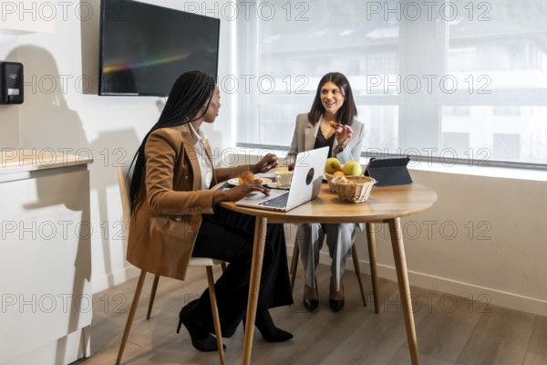Two businesswomen are enjoying breakfast and working on a laptop during their business trip, showcasing the blend of work and leisure in a hotel environment