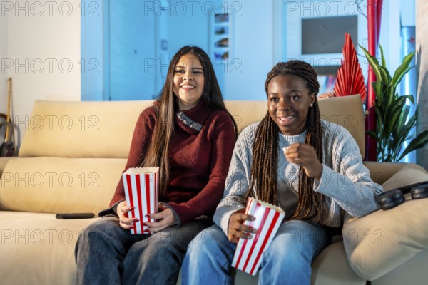 Two happy teenage girls eating popcorn while watching a movie at home, sharing a moment of leisure and friendship