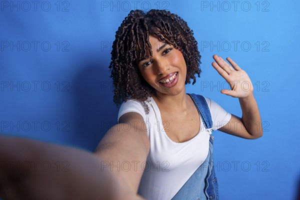 Smiling young woman with curly hair, taking a selfie and waving her hand against a vibrant blue background, radiating happiness and friendliness