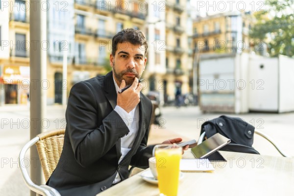Thoughtful businessman gesturing hand on chin while working using mobile phone and digital tablet sitting in an outdoors cafeteria