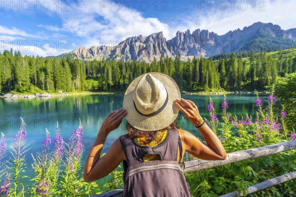 Woman with backpack and straw hat admiring the turquoise waters of lake carezza and the latemar mountain range in the dolomites