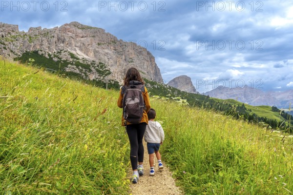 Mother and son walking on a path surrounded by lush green grass, enjoying the breathtaking view of the dolomites mountains in gardena pass