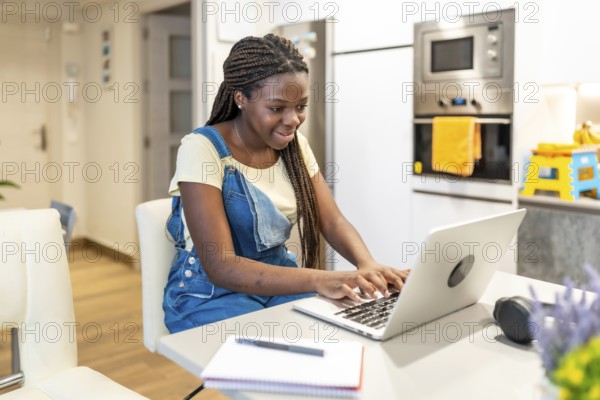 Young student concentrating on her studies, typing on a laptop in a modern kitchen, demonstrating focus and dedication to online learning