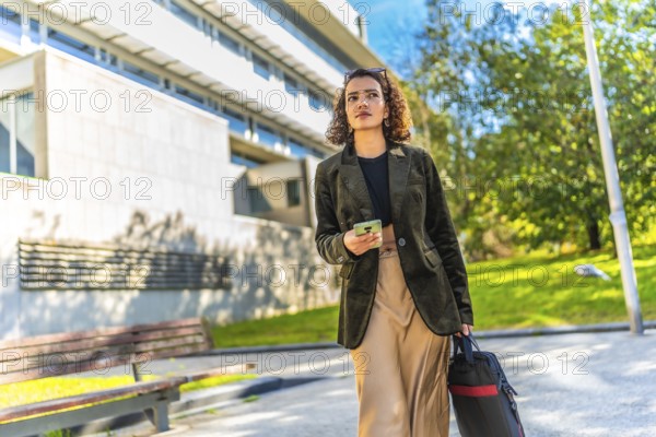 Young arab businesswoman walking along residential area carrying laptop bag and using mobile phone