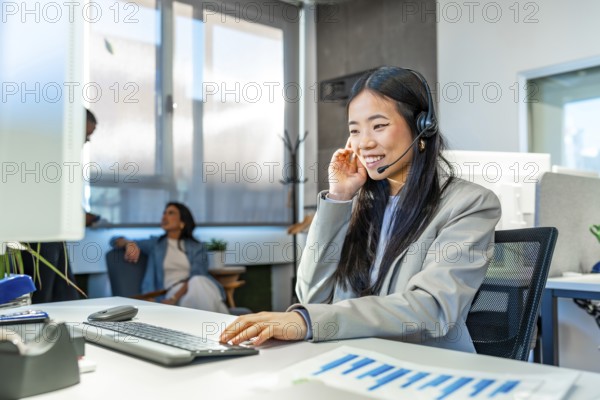 Young asian manager wearing headset working with computer in a coworking office, colleagues talking in the background