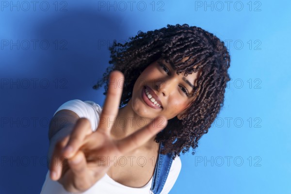 Cheerful young woman smiling and making a peace sign with both hands against a vibrant blue background, radiating joy and positivity