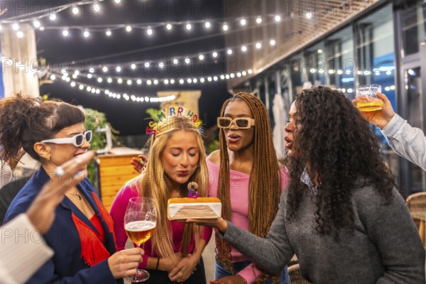 Blonde woman blowing candles of birthday cake celebrating party with friends at night in the terrace of a bar