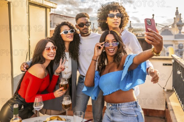 Woman taking selfie with multi-ethnic friends in a roof party with wine and snacks during sunset