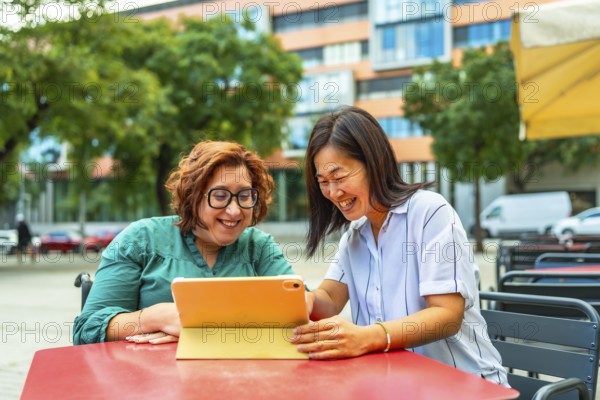 Disabled woman in electric wheelchair and chinese female friend using tablet sitting on sidewalk cafeteria