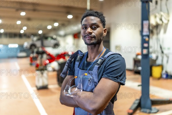 Young african american mechanic standing with crossed arms and protective eyewear in a car repair shop, exuding professionalism