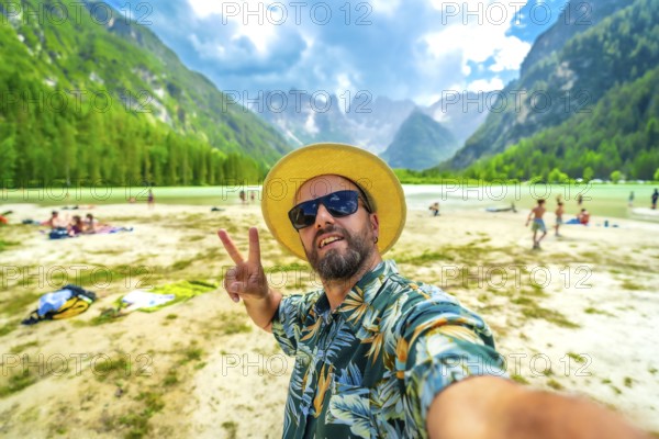Tourist wearing sunglasses and straw hat gesturing peace sign while taking a selfie at the beach of lago di landro in the italian dolomites