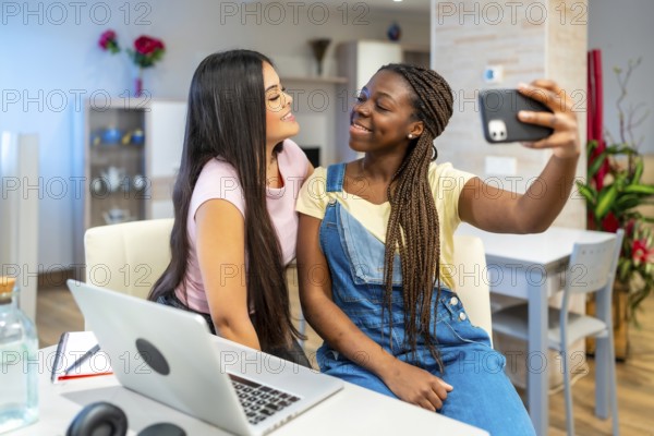 Two happy teenage girls taking a selfie with a smartphone while studying with a laptop at home