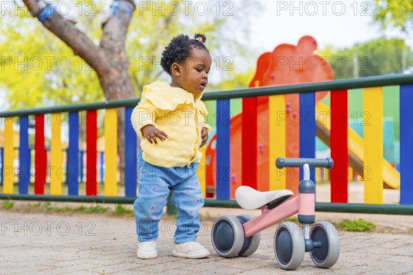 Full length photo of an african baby girl in a playground with a toy bike alone