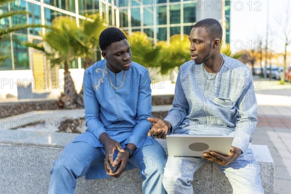 Two senegalese businessmen wearing traditional dashiki attire are sitting outside a modern office building, engaged in a conversation while using a laptop