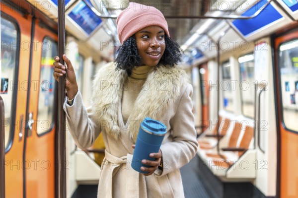 Stylish young woman holding a reusable coffee cup while standing on a subway train, enjoying her commute