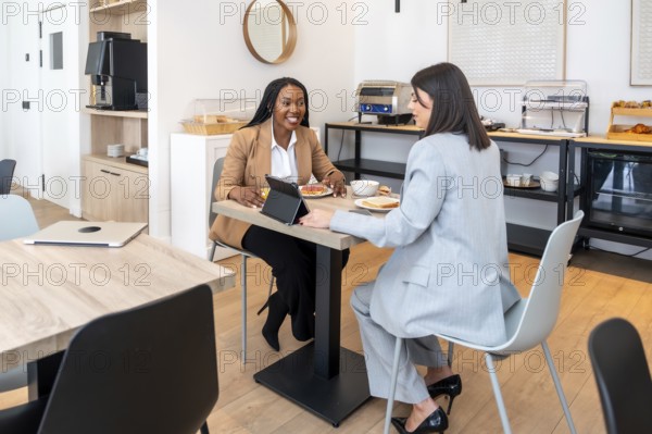 Two businesswomen are enjoying breakfast and discussing work using a digital tablet in a hotel restaurant