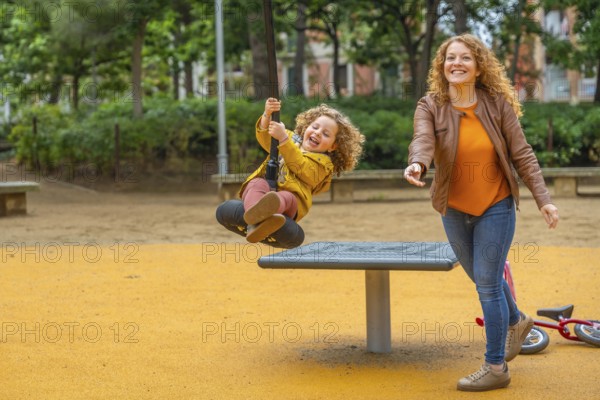 Caucasian adult mother and girl having fun swinging in a playground in an urban park