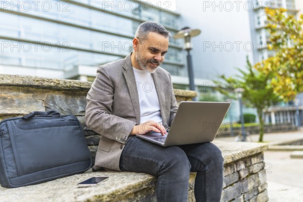 Happy mature caucasian businessman using laptop sitting on a stone bench outdoors in the financial district