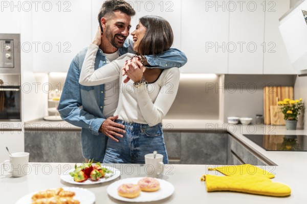Young couple hugging and laughing in their modern kitchen, enjoying a romantic moment together with breakfast ready on the counter