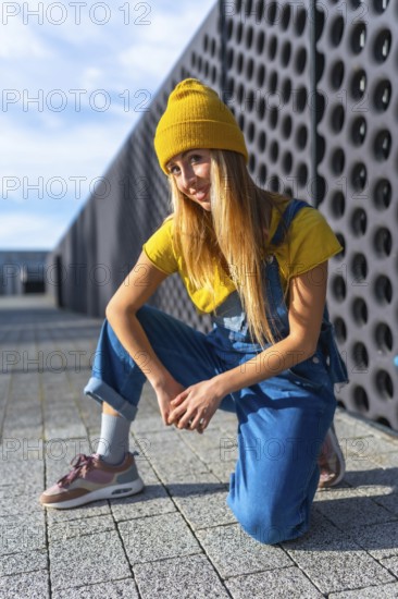 Blonde dancer wearing yellow beanie, yellow t shirt and blue dungarees, kneeling and posing in a modern city setting