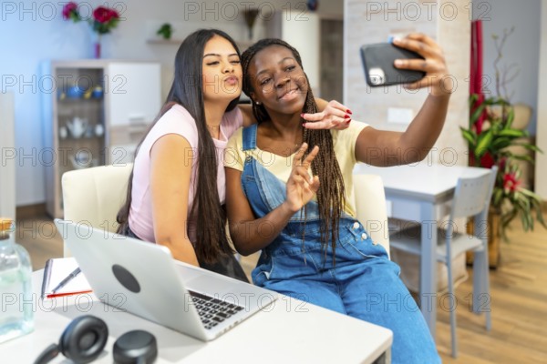 Two friends enjoying a cheerful moment, taking a selfie with a smartphone while studying together at home, embracing their friendship