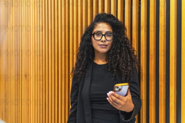 Portrait of an young adult latin businesswoman in black elegant clothes using mobile phone indoors