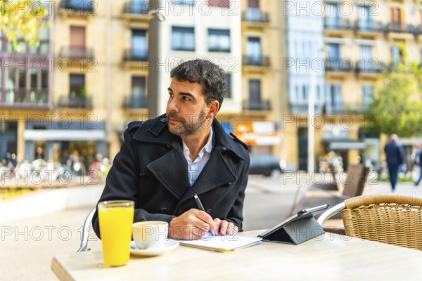 Mature handsome caucasian businessman writing notes and using digital tablet sitting on an outdoors cafeteria