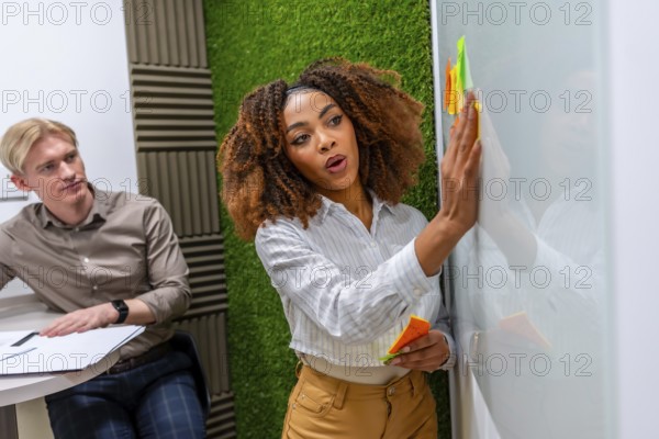 Businesswoman using colorful sticky notes while brainstorming new ideas on a whiteboard in a modern coworking space, with a male colleague sitting nearby