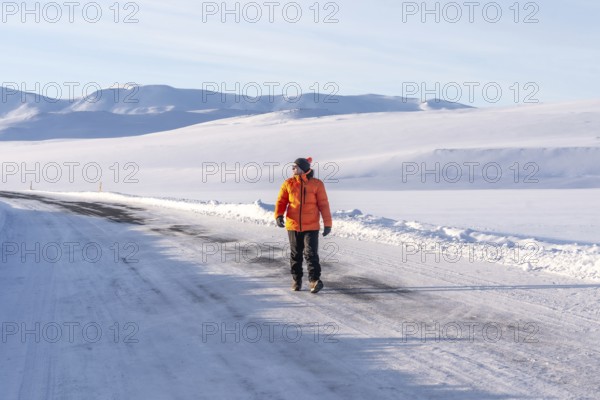 A man in an orange jacket walks across a snow-covered road. The scene is peaceful and serene, with the man being the only person in the image. The orange jacket stands out against the white snow
