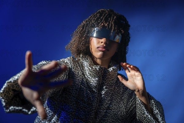Stylish person with futuristic visor and patterned jacket extending hand on vibrant blue background