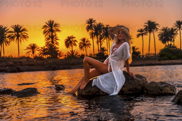 Full length photo of a caucasian woman sitting sensual on water's edge during sunset on a tropical beach