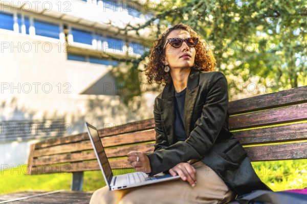 Modern arab businesswoman using laptop sitting on park bench working on remote