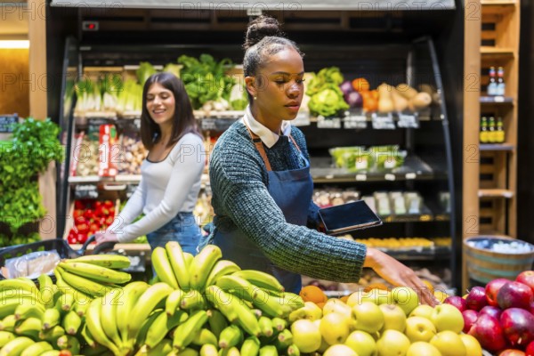 Grocery store employee arranging fresh fruit while holding a tablet, with a customer and shelves full of organic products in background