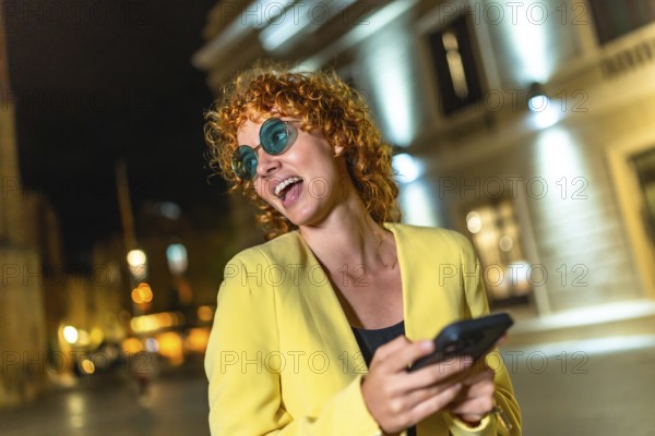 Modern woman in colorful casual clothes and blue sunglasses using smartphone laughing in the city at night
