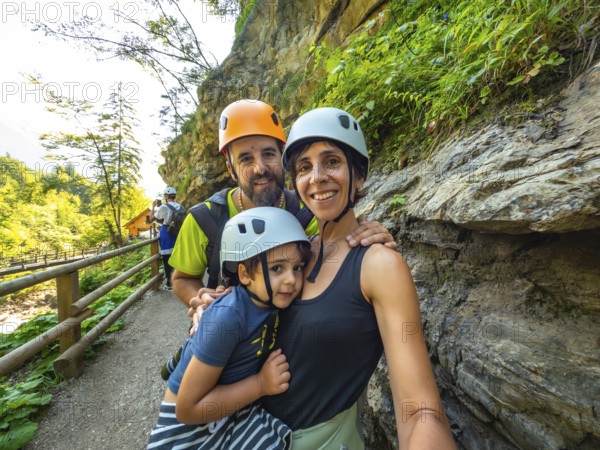 Family wearing helmets taking selfie while hiking on wooden walkway next to rock wall in beautiful vintgar gorge near bled, slovenia