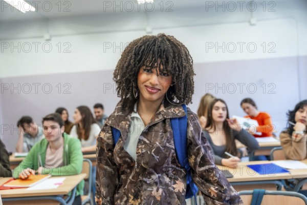 Confident young university student standing in classroom with backpack, classmates studying in background, higher education concept