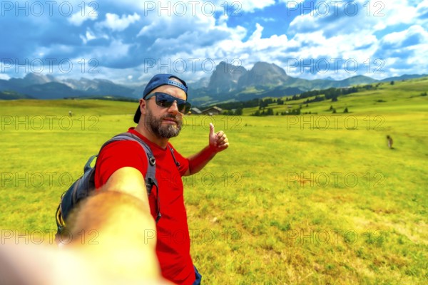 Backpacker taking selfie showing thumbs up sign enjoying beautiful summer landscape at alpe di siusi in dolomites, italy