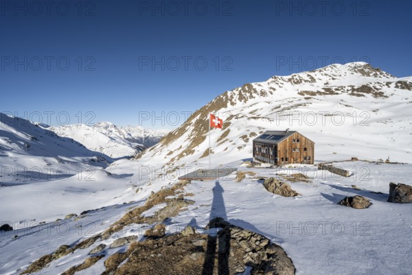 Swiss flag and mountain hut Keschhütte in front of mountain landscape in winter, behind summit of the Bergüner Stöcke Tinzenhorn and Piz Ela, Bündner Haute Route, Albula Alps, Rhaetian Alps, Grisons, Eastern Switzerland, Switzerland, Europe
