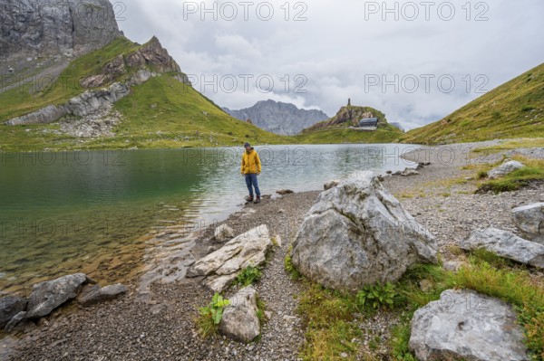 Mountaineer in yellow jacket at the mountain lake, view over the Wolayersee lake to the alpine club hut Wolayerseehütte, cloudy mountain landscape, Carnic Alps, Carnic High Trail, Carinthia, Austria, Europe