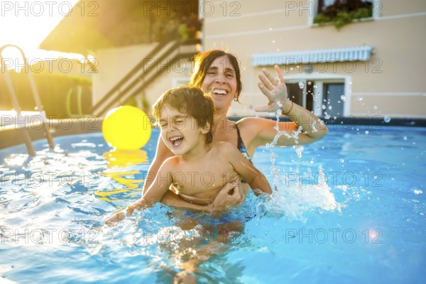 Happy mother and son are having fun, splashing and playing in a swimming pool during summer vacation at sunset