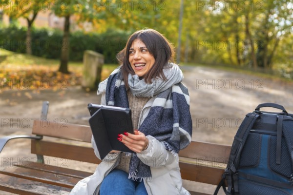 Happy caucasian female student using digital tablet sitting on a park bench during a cold day of autumn