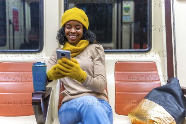 Smiling young woman wearing yellow gloves and beanie using smartphone while commuting on subway train