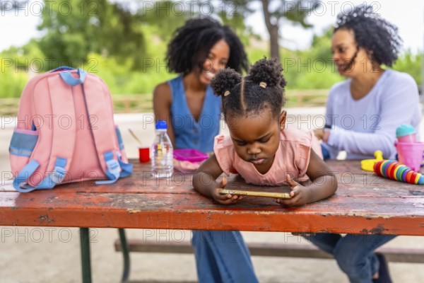 Little african girl using mobile phone in the park with her grandmother and mother talking on the background