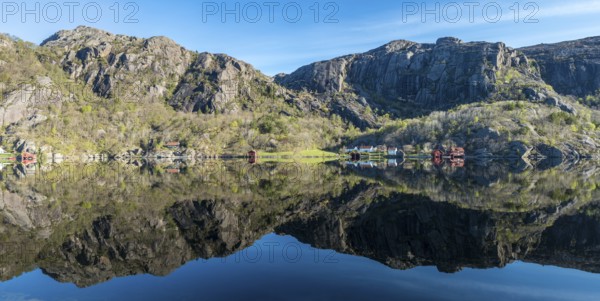 Near Ana-Sira at the norwegian southern coast, lonely houses at the foot of a rock formation are reflected in the calm sea, panorama, Norway, Europe