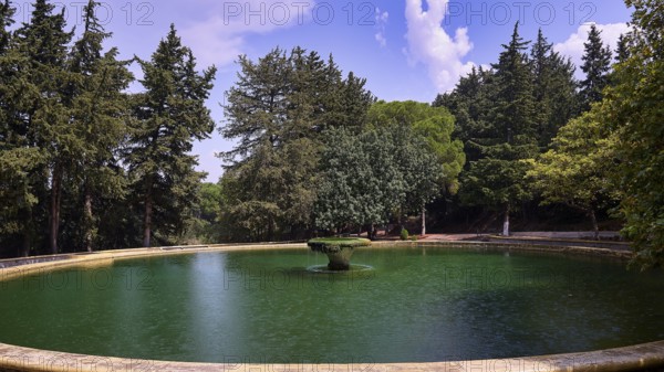 A round lake with a central fountain surrounded by tall green trees under a blue sky with clouds, Italian Governor's Palace, Ruins, Italian colonial settlement, Eleousa, Rhodes, Dodecanese, Greek Islands, Greece, Europe