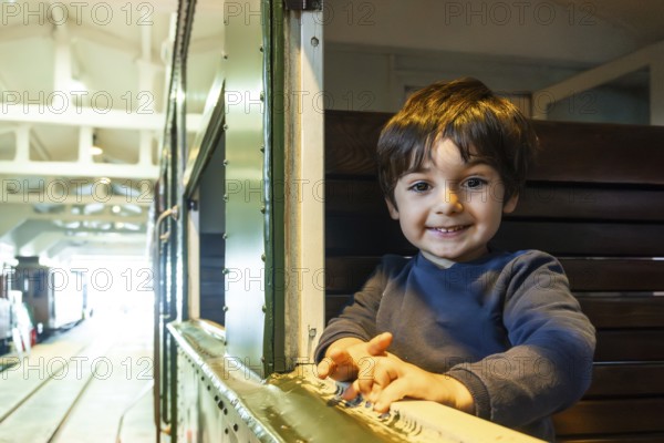 A young boy is smiling and looking out a window of a train. The train is in a station and the boy is sitting on a bench