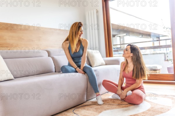 Two smiling female friends in sportswear chatting and relaxing after practicing yoga together in their modern apartment living room