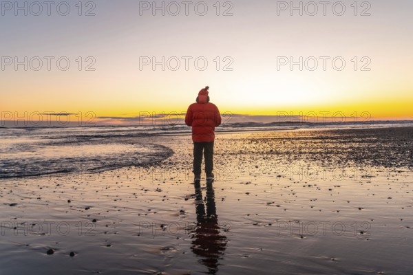 A man stands on a beach at sunset, wearing a red jacket. The scene is serene and peaceful, with the man's reflection in the water adding to the calm atmosphere