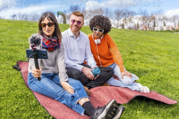 Three content creators sitting on a blanket in a park, recording a video blog using a smartphone with microphone